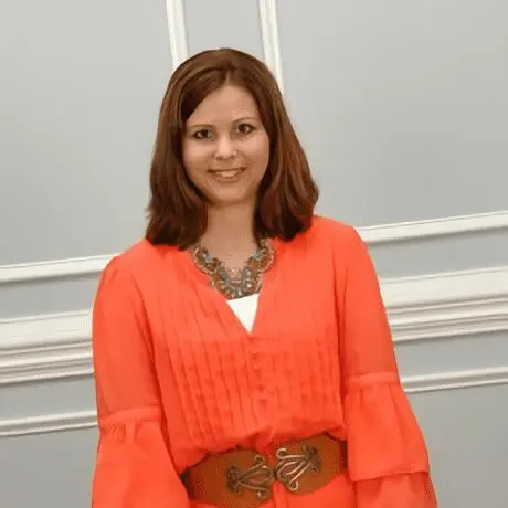 A woman in an orange blouse smiling indoors against a gray wall.