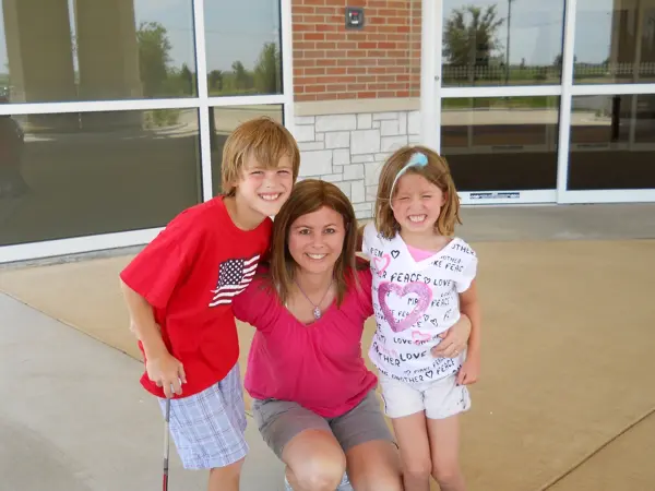 Smiling woman crouches with two happy children outside a building.