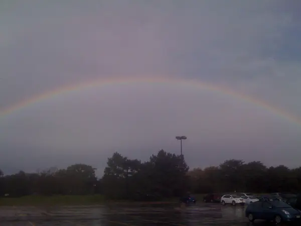 A subtle rainbow arches over a cloudy sky above trees and parked cars.