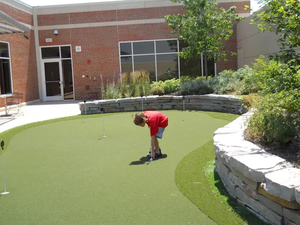 A boy playing mini-golf on a green putting surface near a brick building.