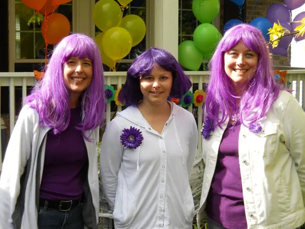 Three women with purple wigs and white outfits smiling indoors.