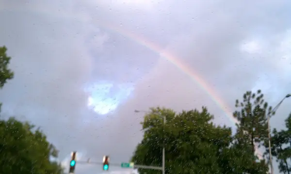 A vibrant rainbow arches across a cloudy sky above traffic lights and trees.