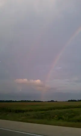 A faint rainbow arches across a cloudy sky above a flat green field.