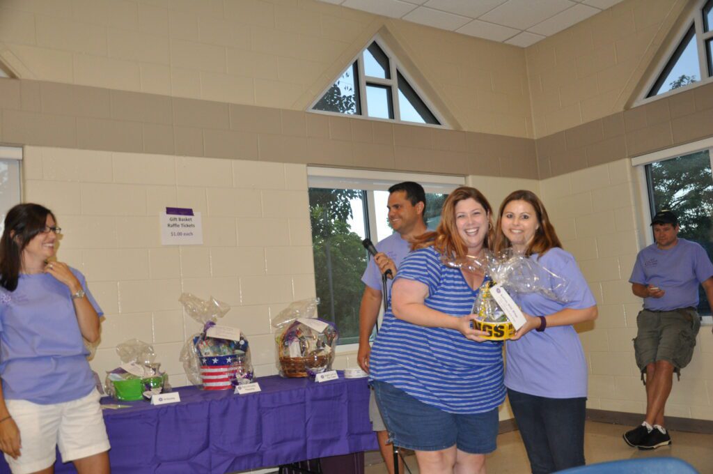 Three people smiling and holding a basket at an indoor event with a table of items.