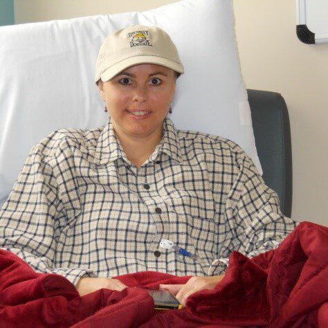 A smiling woman in a hospital bed wearing a cap and plaid shirt.